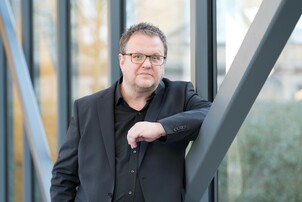 Ein Mann mit kurzen haaren und Brille lehnt an einem schrägen Metallträger. 