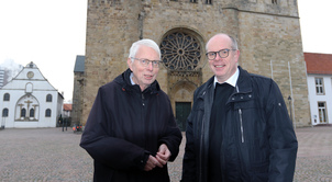 Zwei dunkel gekleidete Männer stehen vor einer Kirche.  